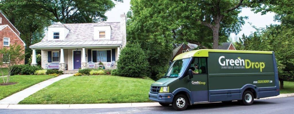 A GreenDrop donation collection van is parked on a suburban street in front of a house with a porch and lawn, surrounded by trees and greenery.