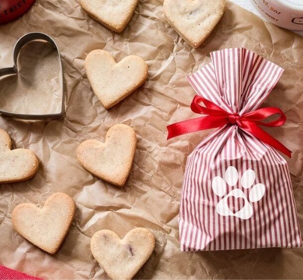 Heart-shaped homemade dog biscuits and a metal cookie cutter rest on brown parchment paper beside a red-and-white striped gift bag tied with a red ribbon and decorated with a white paw print. A red ribbon and pink cloth complete the scene.