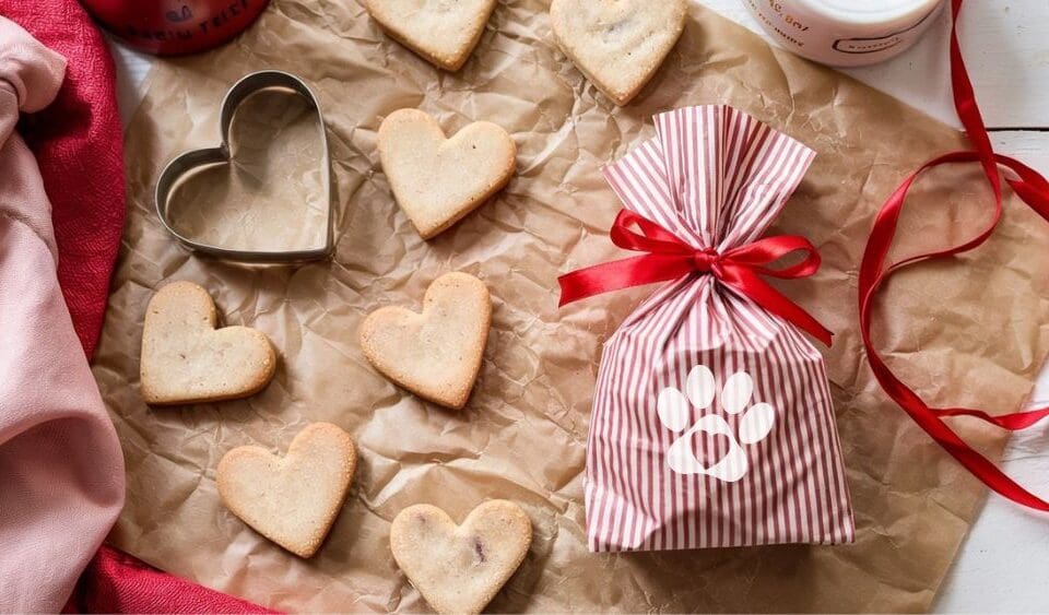 Heart-shaped homemade dog biscuits and a metal cookie cutter rest on brown parchment paper beside a red-and-white striped gift bag tied with a red ribbon and decorated with a white paw print. A red ribbon and pink cloth complete the scene.