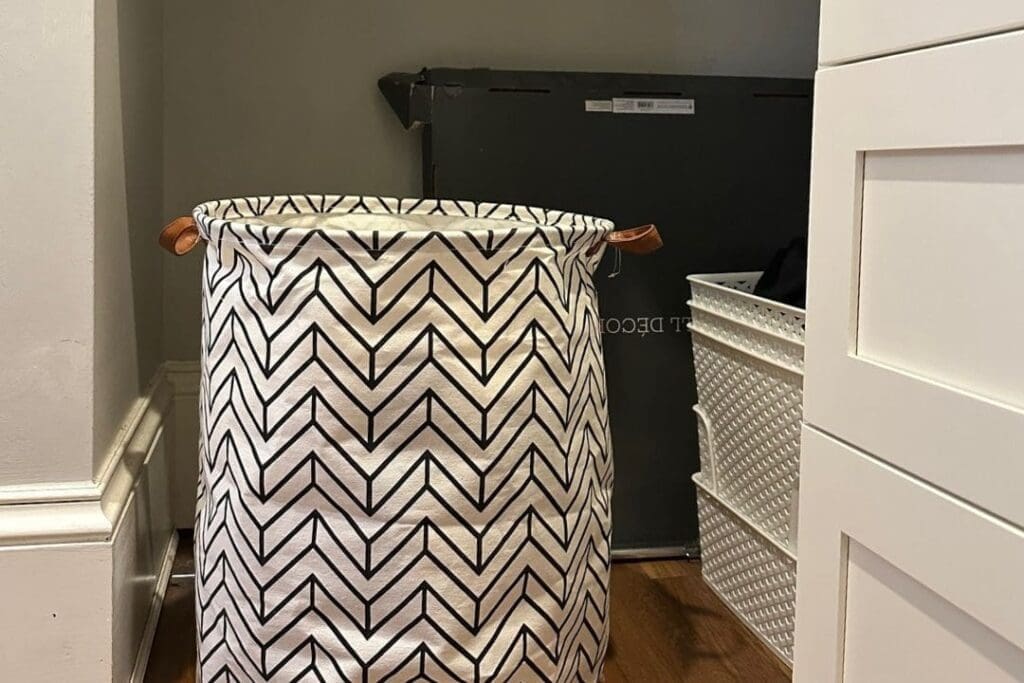 A white laundry hamper with a black zigzag pattern and brown handles sits on a wooden floor in a small corner next to a white dresser and a plastic basket. A dark object leans against the wall in the background.