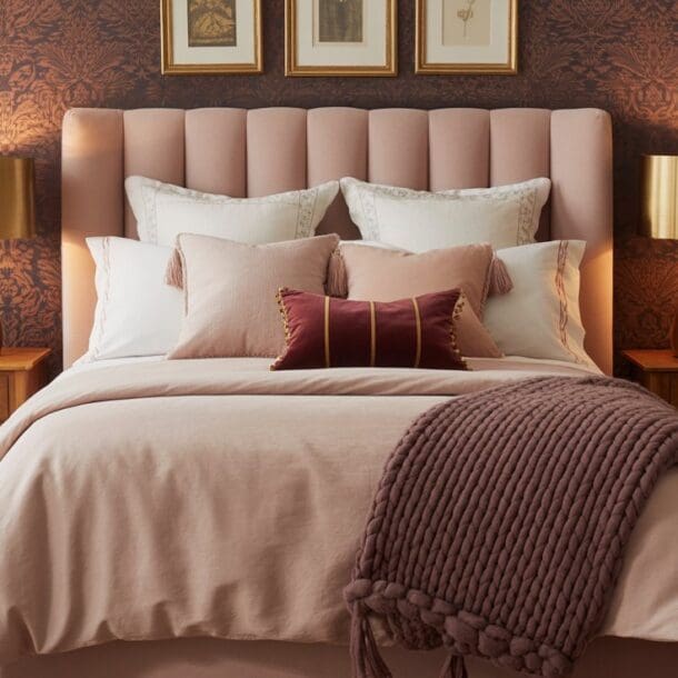 A neatly made pink and burgundy bedroom features a blush pink tufted headboard, gold lamps on wooden nightstands, white and pink pillows, a mauve throw blanket, and dark patterned wallpaper with framed art above the bed.