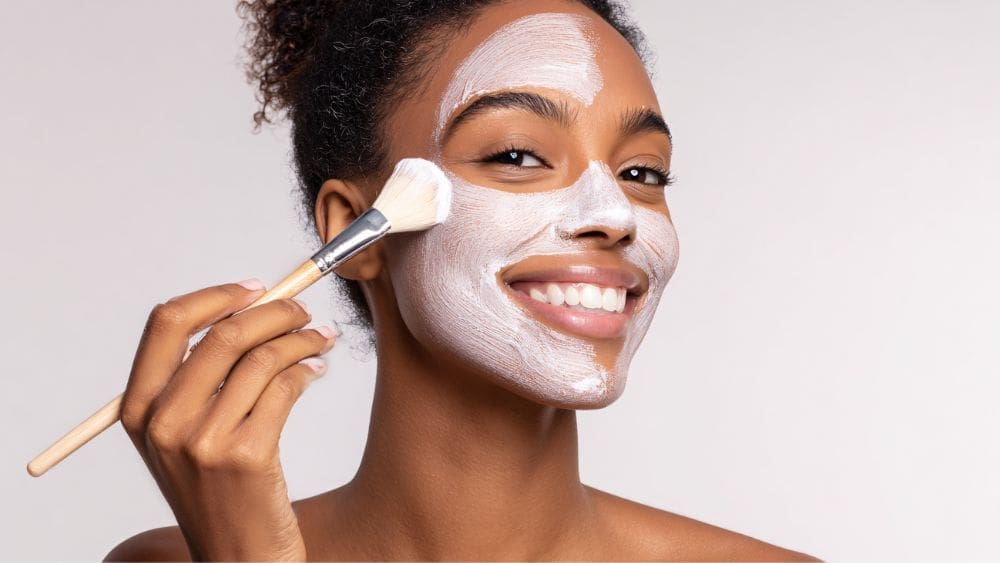 A smiling woman applies a white facial mask to her face with a brush, against a plain light background.