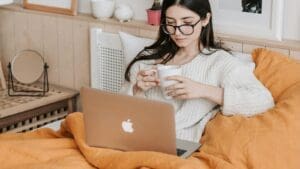 A woman with long dark hair and glasses sits in bed under an orange blanket, holding a white mug and looking at a laptop. She appears relaxed, with cozy bedroom decor in the background.