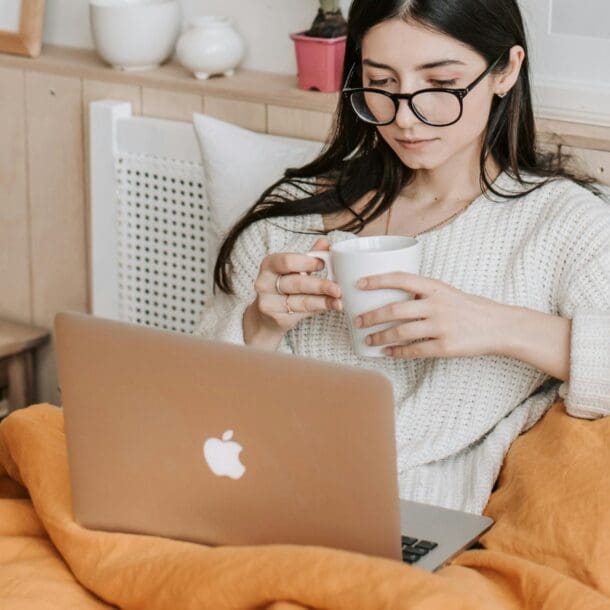 A woman with long dark hair and glasses sits in bed under an orange blanket, holding a white mug and looking at a laptop. She appears relaxed, with cozy bedroom decor in the background.