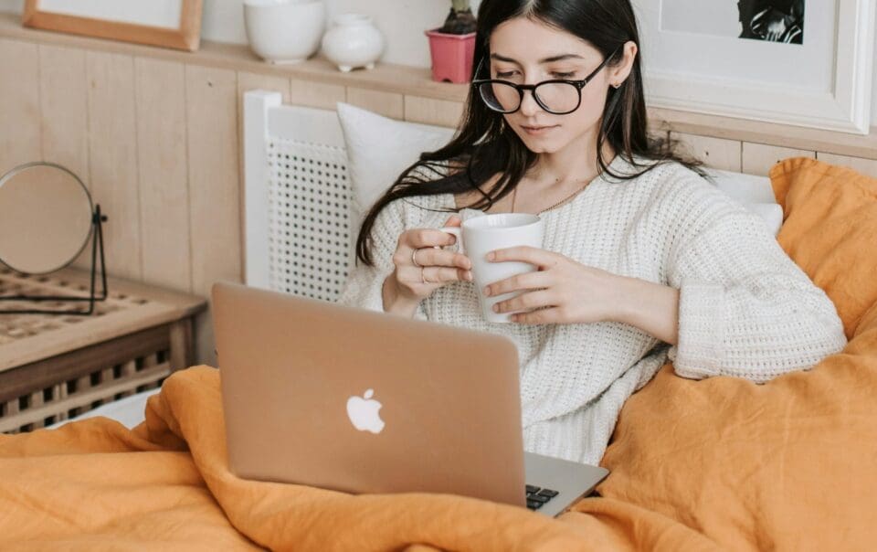 A woman with long dark hair and glasses sits in bed under an orange blanket, holding a white mug and looking at a laptop. She appears relaxed, with cozy bedroom decor in the background.