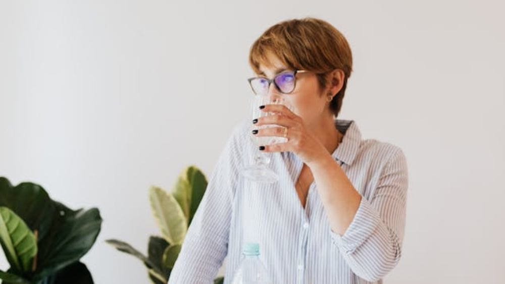 A person with short hair and glasses, wearing a striped shirt, drinks from a clear glass. Green potted plants are in the background against a plain white wall.