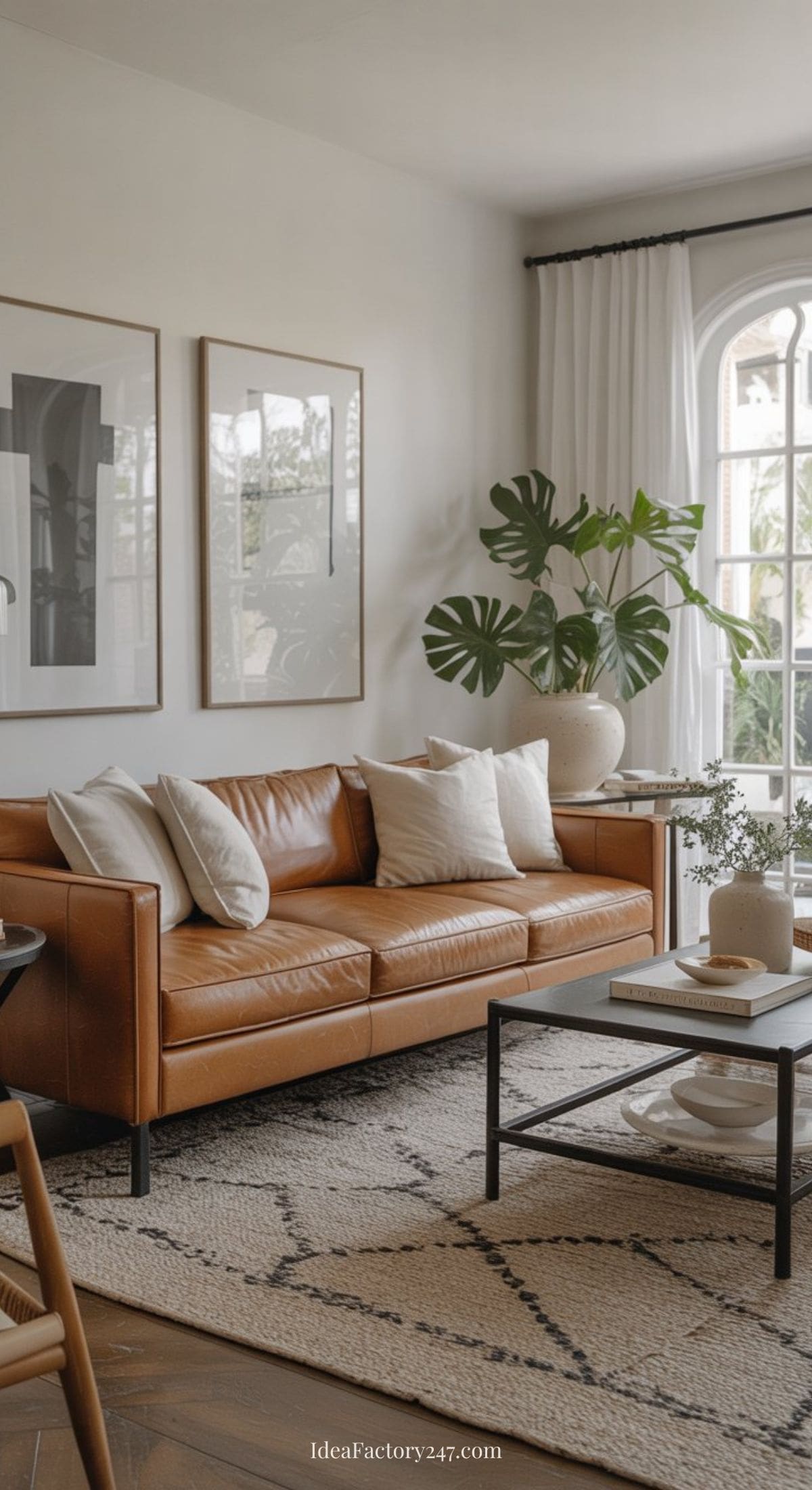 A modern living room with a tan leather sofa, white cushions, a patterned rug, framed wall art, a coffee table with decor, a large potted plant, and sunlight streaming through a tall arched window with white curtains.
