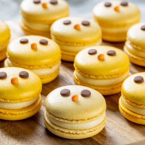A group of yellow macarons decorated to look like chicks, with small chocolate eyes and orange candy beaks, arranged on a wooden platter.