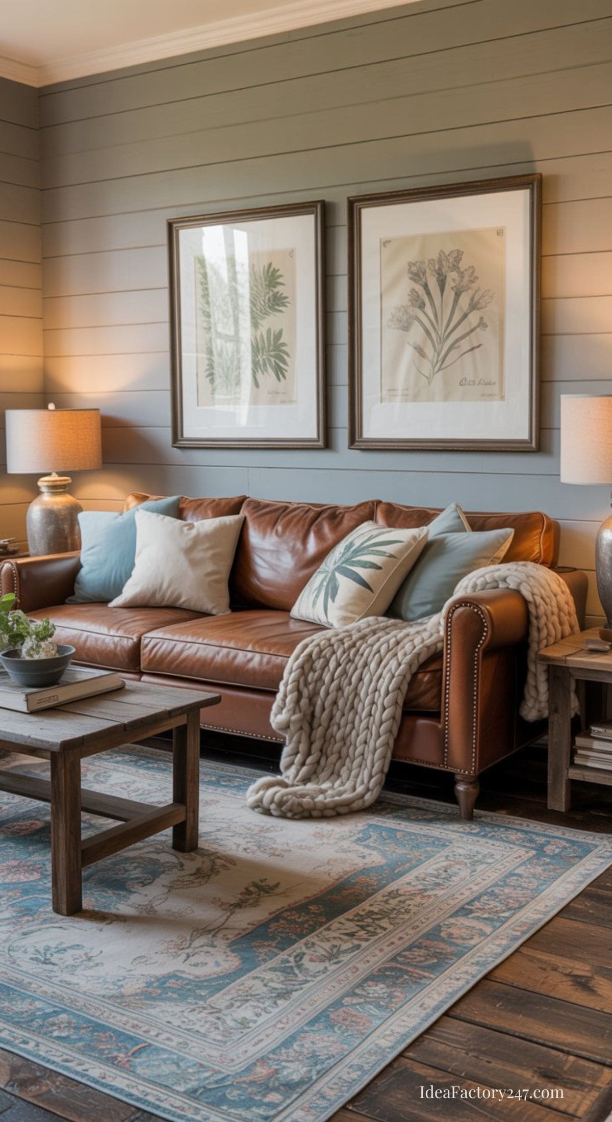 A cozy living room with a brown leather sofa, light blue and beige pillows, a chunky knit throw, two wooden coffee tables, matching table lamps, botanical artwork, and a patterned rug on a wooden floor.