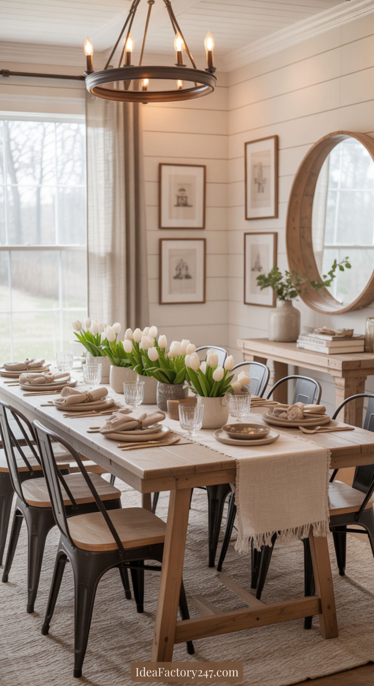 A bright dining room with a long wooden table set for a meal, decorated with white tulips in vases. Black chairs surround the table, and a round mirror, framed art, and chandelier add to the cozy, elegant decor.