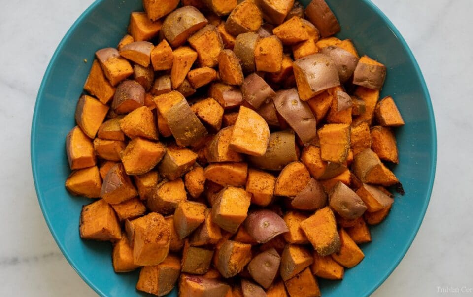 A blue bowl filled with seasoned, cubed sweet potatoes with cinnamon sits on a white marble surface.