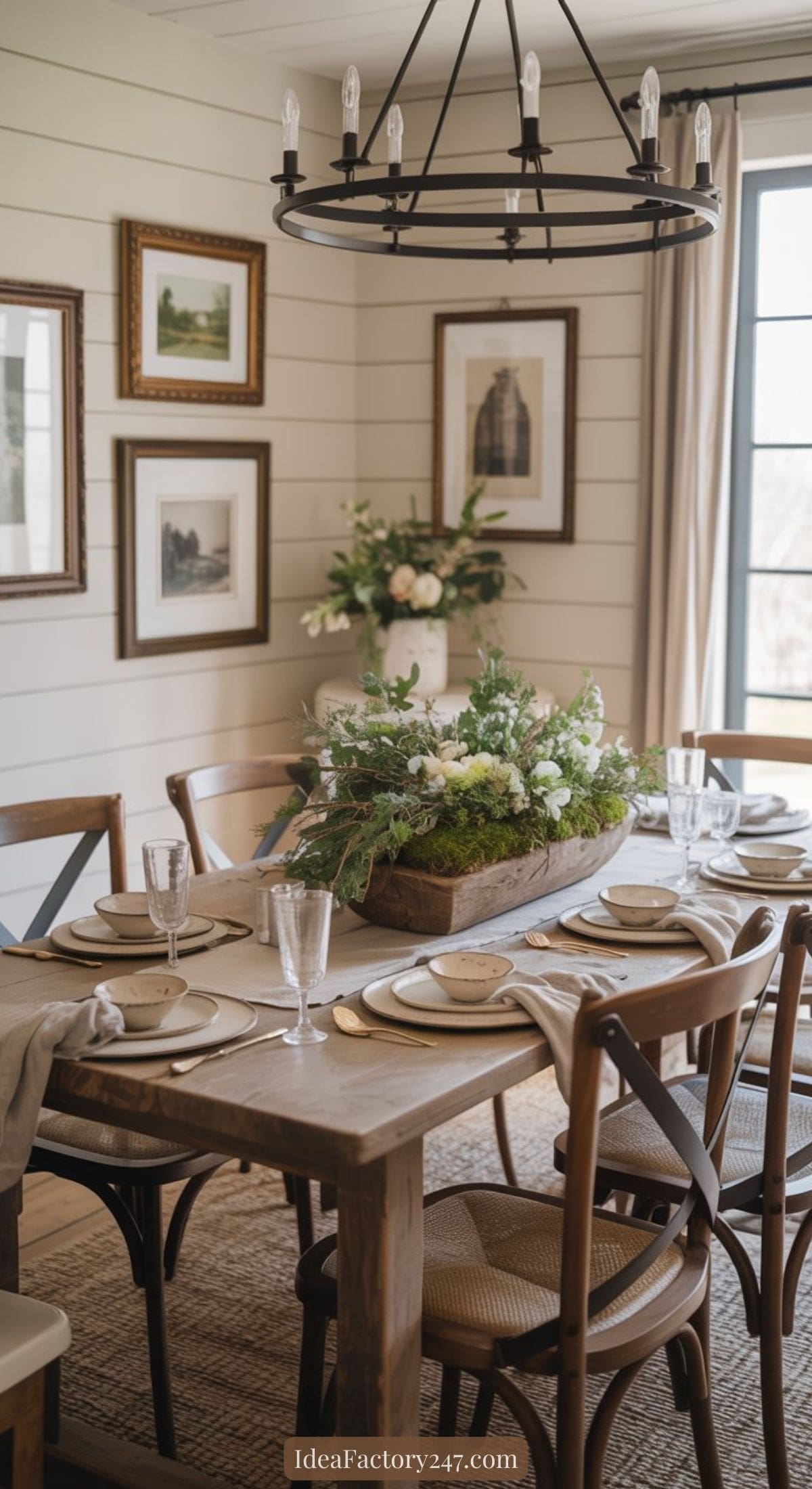 A rustic dining room with a wooden table set for six, featuring neutral plates and glassware. A centerpiece of greenery and white flowers decorates the table. Framed art and a black chandelier add charm to the bright, airy space.