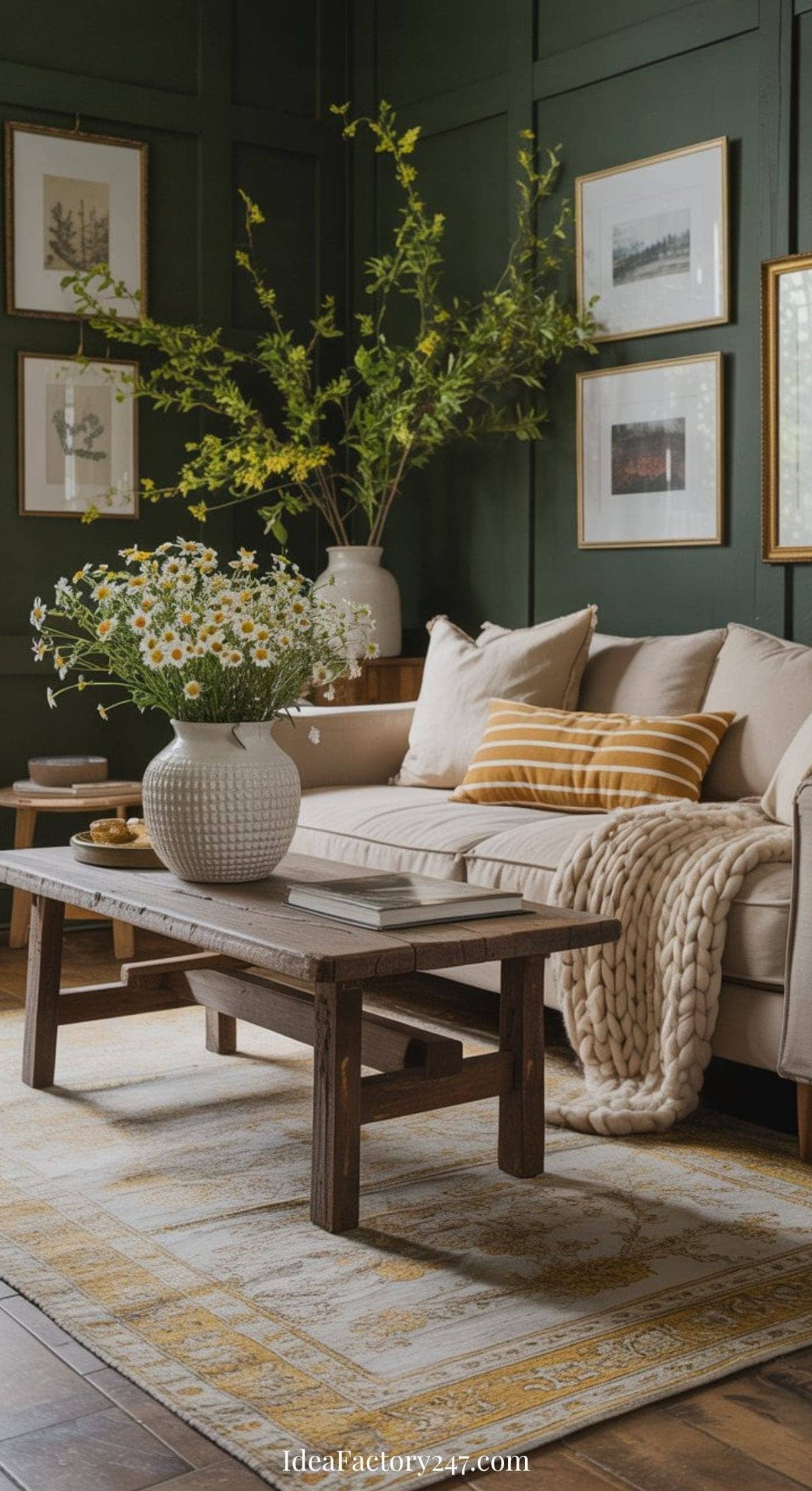 A cozy living room with a beige sofa, yellow striped pillow, chunky knit blanket, wooden coffee table with a white vase of daisies, green paneled walls, framed art, and abundant plants.