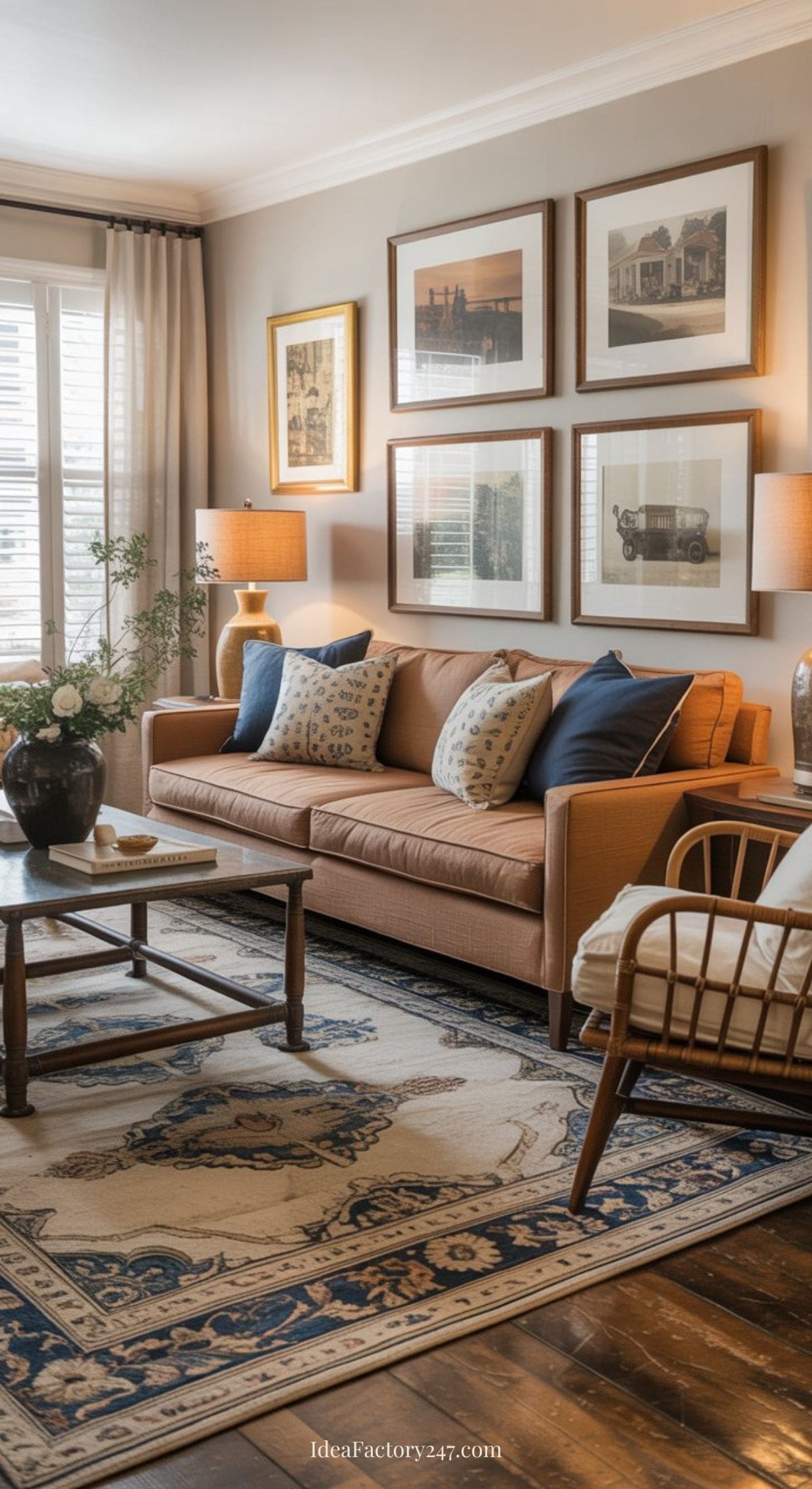 A cozy living room with a brown couch, blue and patterned pillows, a wooden coffee table, framed vintage art on the wall, two table lamps, a woven chair, and a patterned rug over hardwood floors.