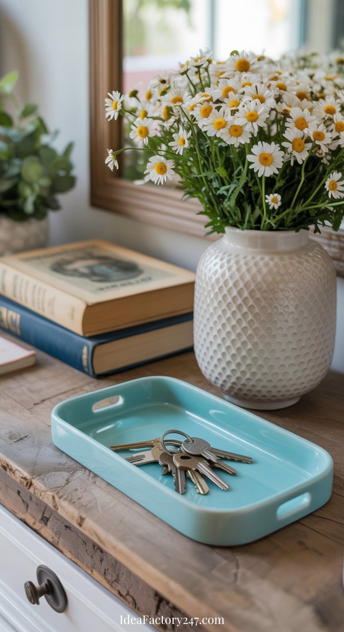 A light blue tray with keys sits on a wooden table, styled with spring entryway decor—a stack of books and a white vase filled with daisies. A mirror and green plant complete the fresh, welcoming scene.
