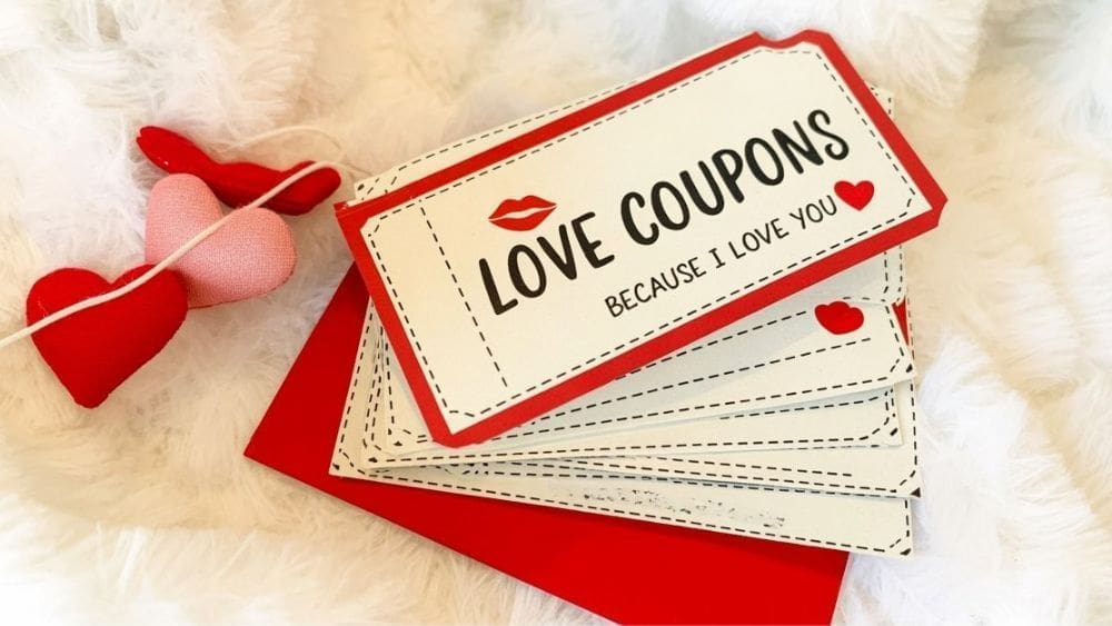 A stack of Love Coupons with red borders and heart designs, placed on a red envelope atop a fluffy white surface. Three fabric hearts are visible beside the coupons, making them perfect as Thoughtful Valentine’s Gifts.