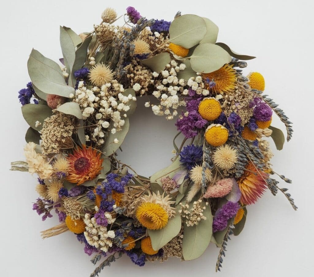 A wreath made of various dried flowers and leaves, featuring shades of yellow, orange, white, purple, and beige, arranged in a circular shape against a plain white background.