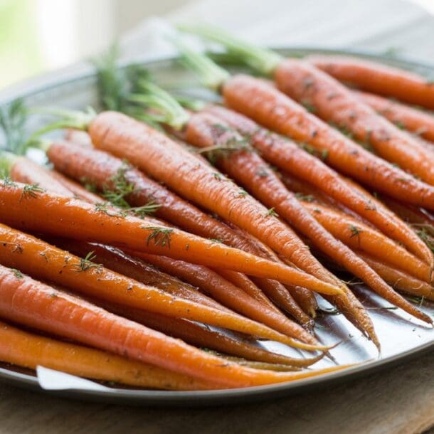 A silver tray filled with fresh, whole carrots, some with green tops still attached, sits on a rustic wooden surface. The neatly arranged carrots are sprinkled with herbs, ready to be transformed into honey roasted carrots.