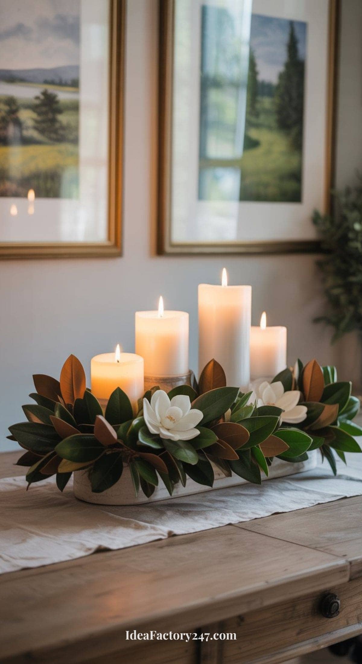 A centerpiece features four lit white pillar candles surrounded by green leaves and white flowers, arranged on a wooden table with a white runner. Framed landscape photos hang on the wall in the background.