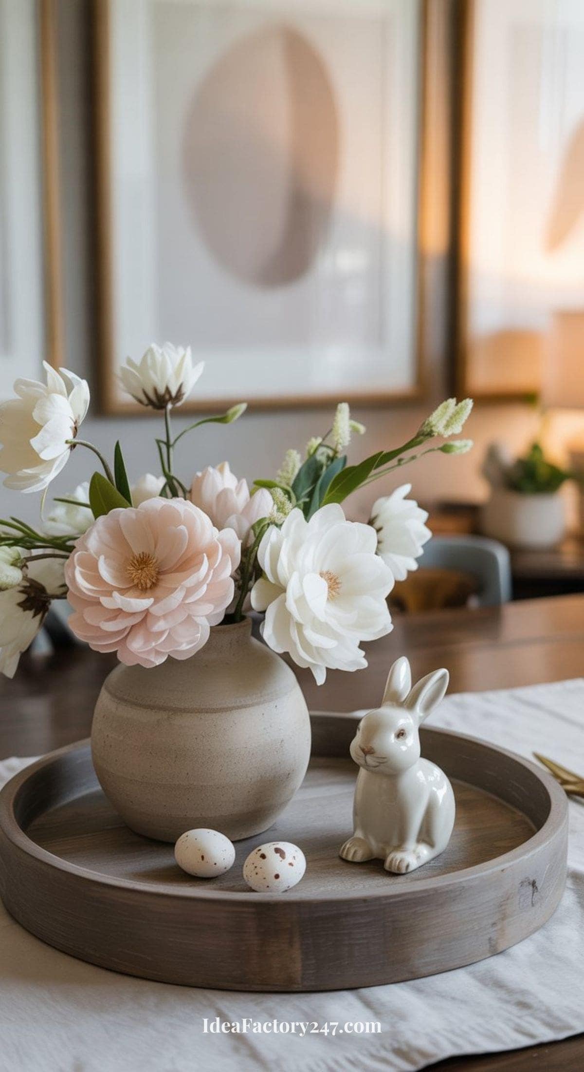 A wooden tray on a table holds a ceramic vase with white and pale pink flowers, a small ceramic bunny figurine, and two speckled eggs. Framed abstract art is blurred in the background.