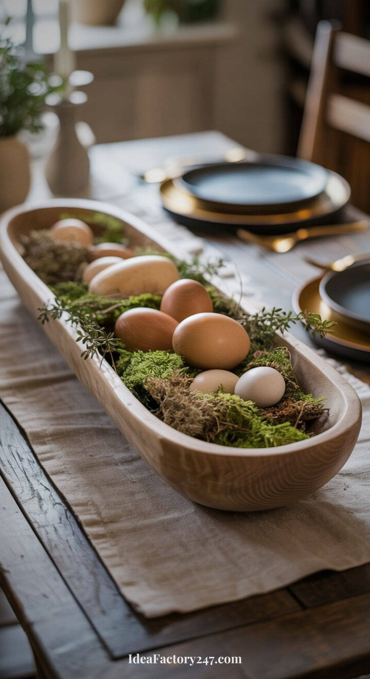 A wooden bowl filled with brown and white eggs, moss, and greenery sits on a linen table runner as a centerpiece on a rustic dining table set with plates and gold-rimmed chargers.