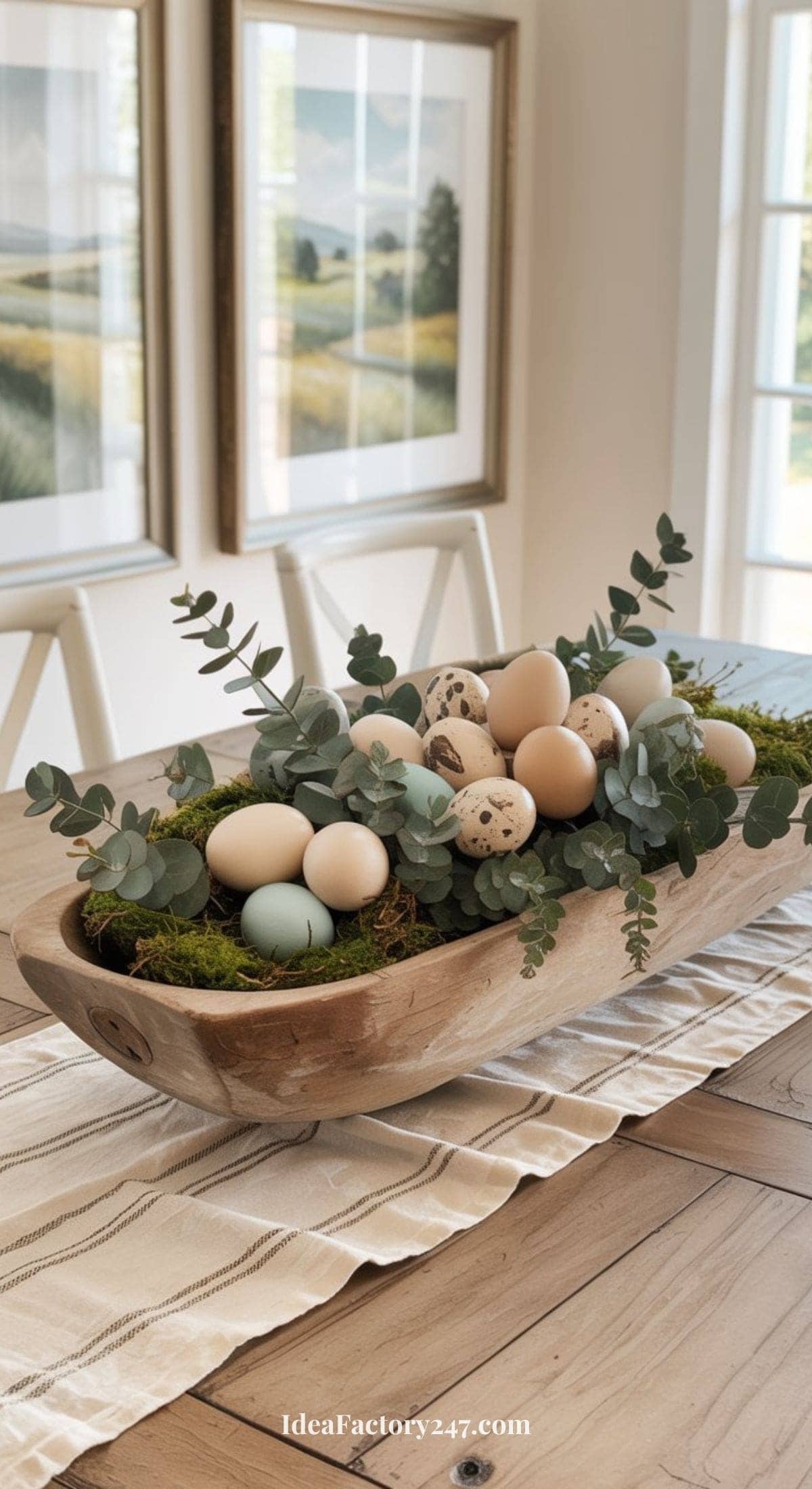 A wooden bowl filled with moss, greenery, and pastel-colored eggs sits on a striped table runner atop a wooden dining table, with framed landscape art and sunlight in the background.