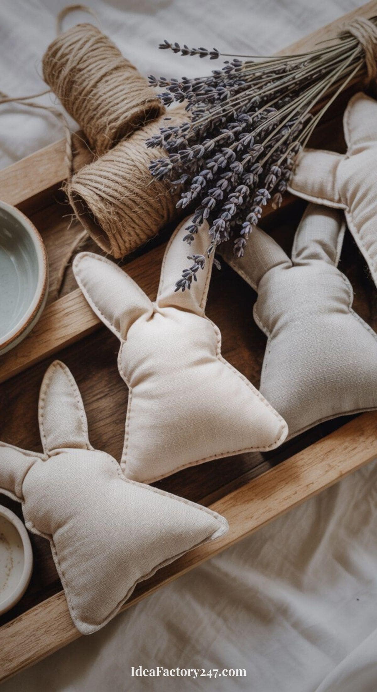 A wooden tray holding three fabric bunny decorations, a bundle of dried lavender, twine, and a ceramic bowl, all arranged on a soft, neutral-colored cloth.