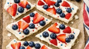 Rectangular pieces of dessert bars topped with white frosting, fresh blueberries, sliced strawberries, and red and blue sprinkles, arranged on parchment paper. A striped cloth is partially visible on the side.