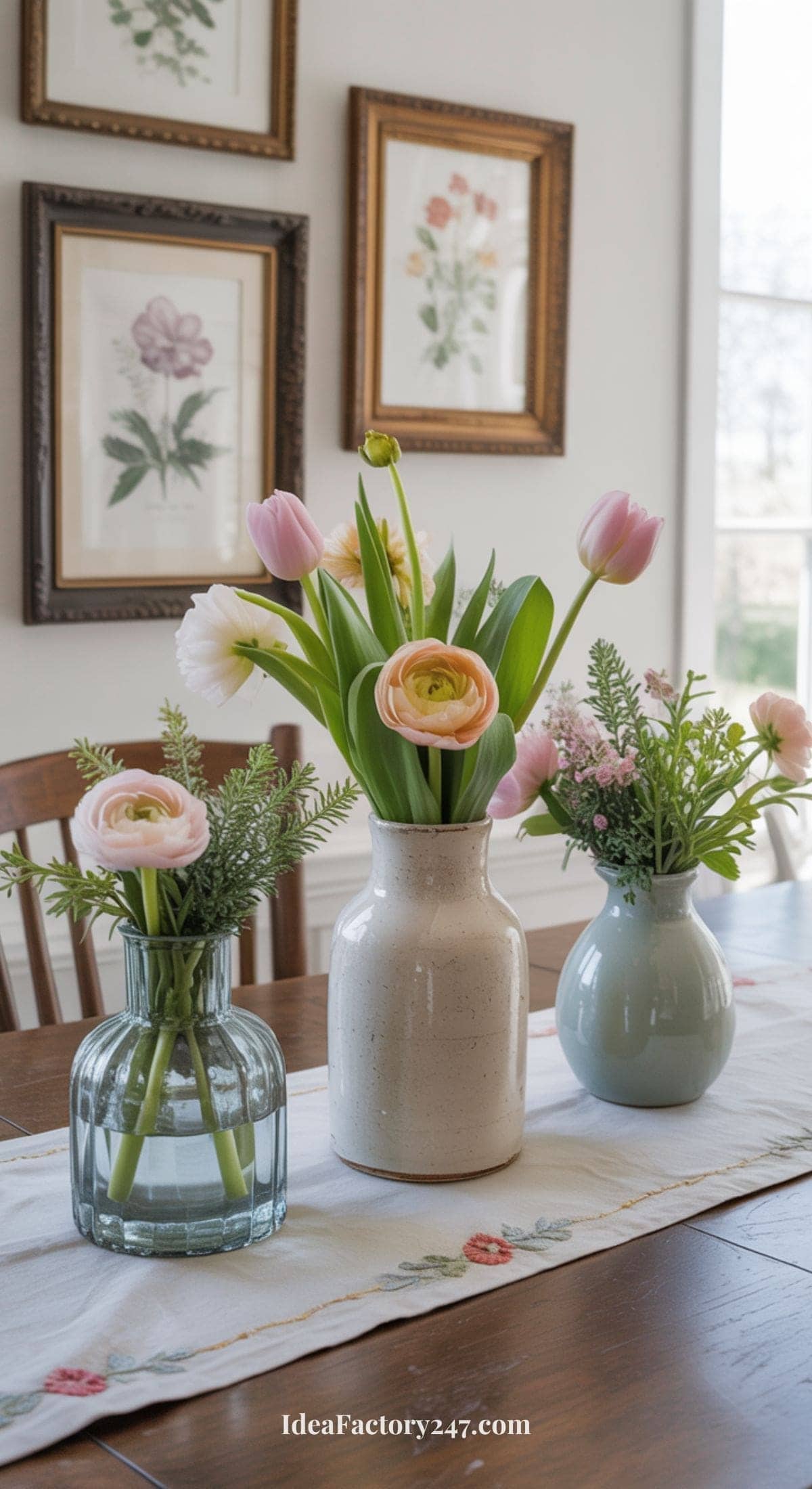 Three vases with assorted pink and white flowers sit on a table runner in a bright dining room. Framed botanical prints hang on the wall behind the arrangement.