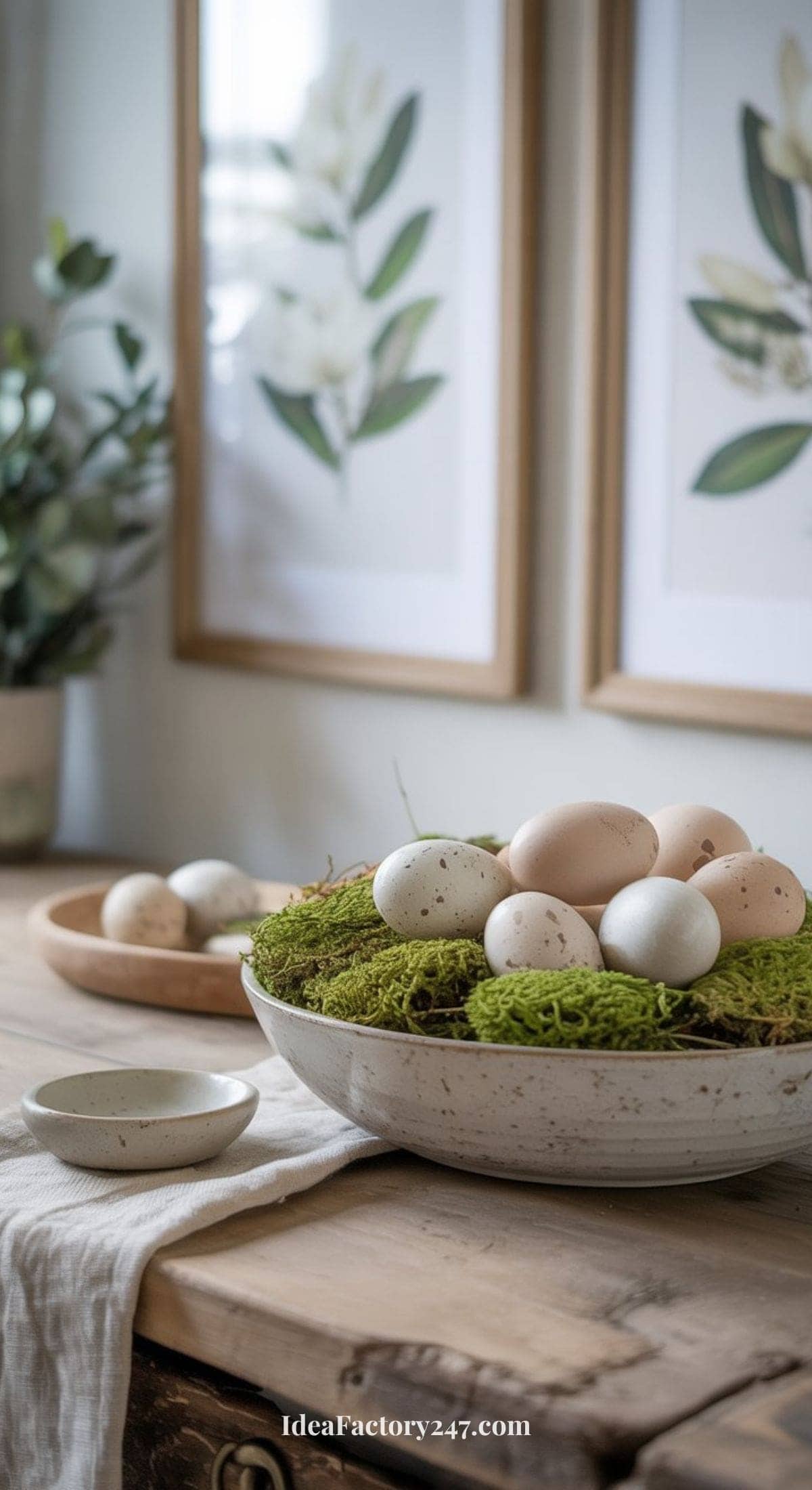 A ceramic bowl filled with moss and speckled eggs sits on a rustic wooden table. In the background are two botanical prints and a leafy plant, creating a natural, spring-inspired setting.