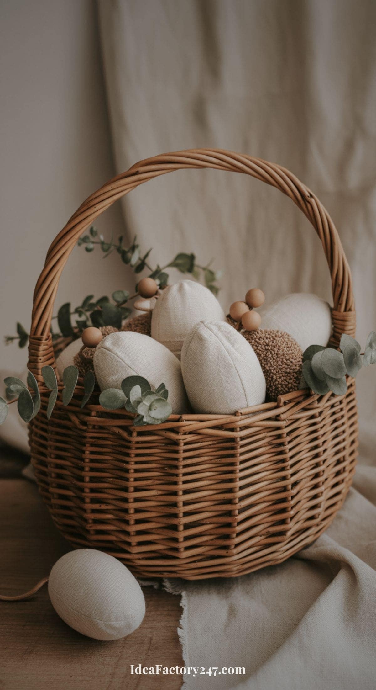 A wicker basket filled with fabric eggs, greenery sprigs, and wooden beads sits on a wooden surface with a neutral fabric backdrop. One fabric egg rests outside the basket.