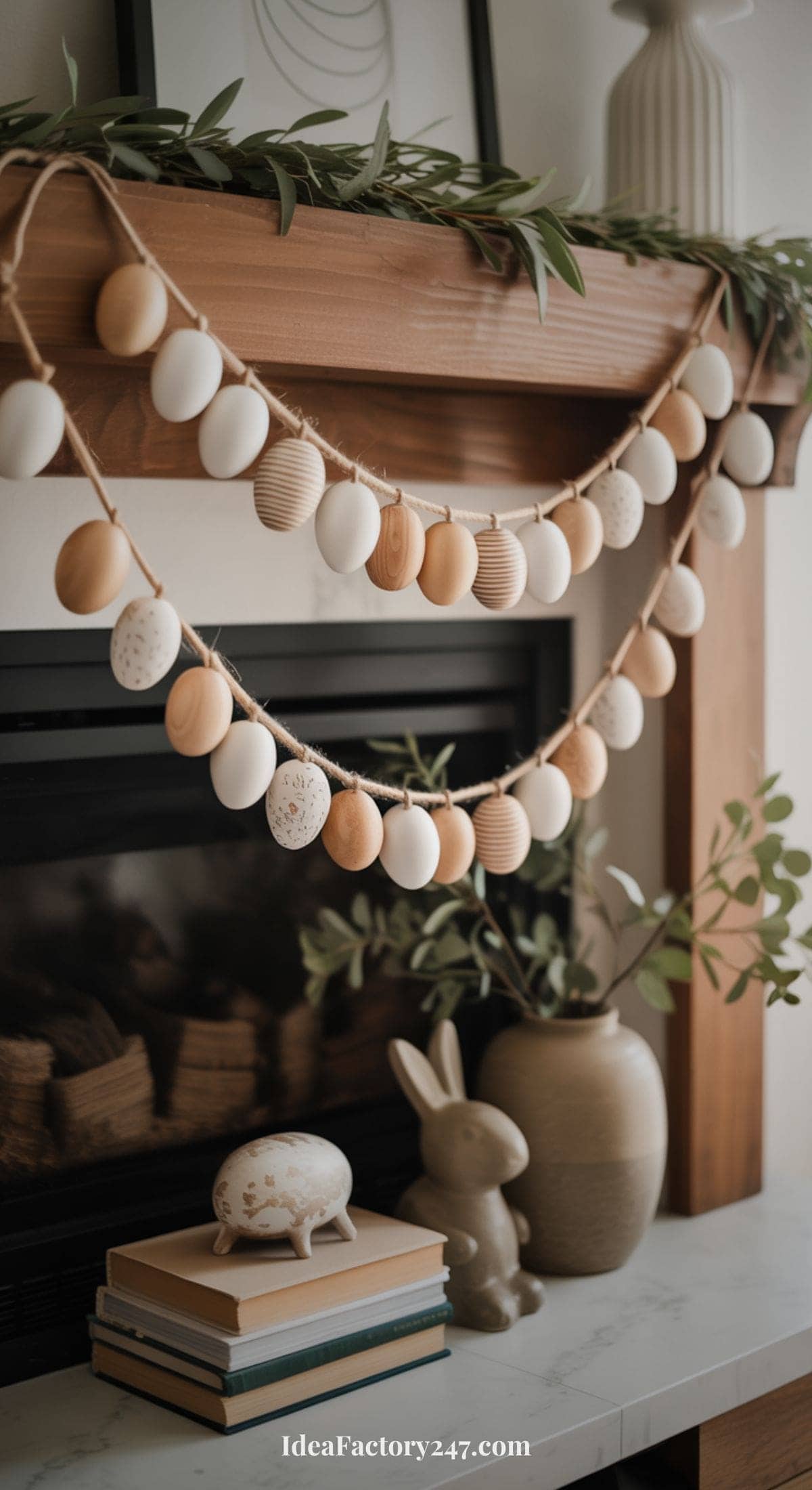 Two garlands of wooden and white painted eggs hang on a wooden mantel. Below, a ceramic bunny, a decorative egg, and stacked books sit beside a vase with green branches. The mantel is decorated with greenery and framed art.