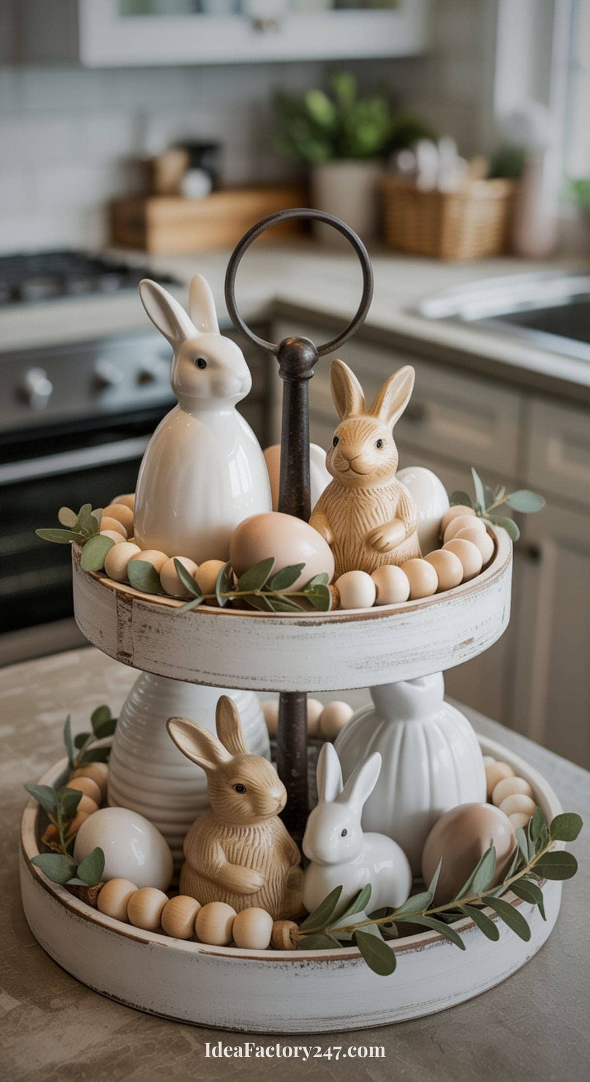 A two-tiered white tray holds ceramic bunnies, eggs, and greenery, creating a festive Easter display on a kitchen counter. The background shows a kitchen with cabinets, a stove, and decor items.