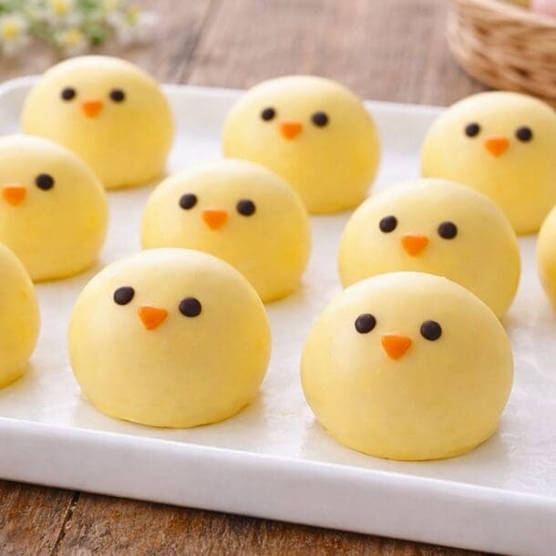 A tray of No Bake Chick Cheesecake Bites—round yellow chick-shaped sweets with chocolate eyes and orange beaks—are arranged neatly on white parchment paper, with pastel candy eggs in the background.