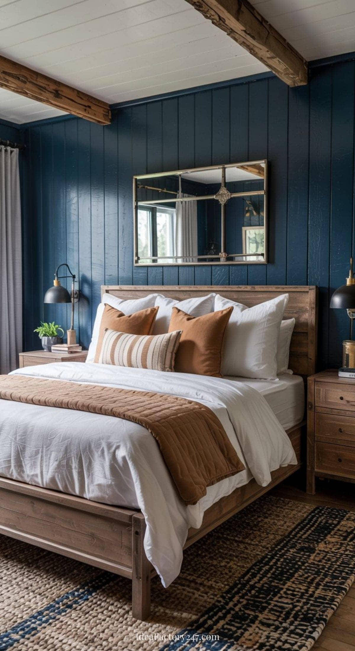 A cozy farmhouse bedroom features a wooden bed with white and brown bedding, blue paneled walls, a large mirror, bedside tables with lamps, and a woven rug on the floor. Wooden beams run across the white ceiling.