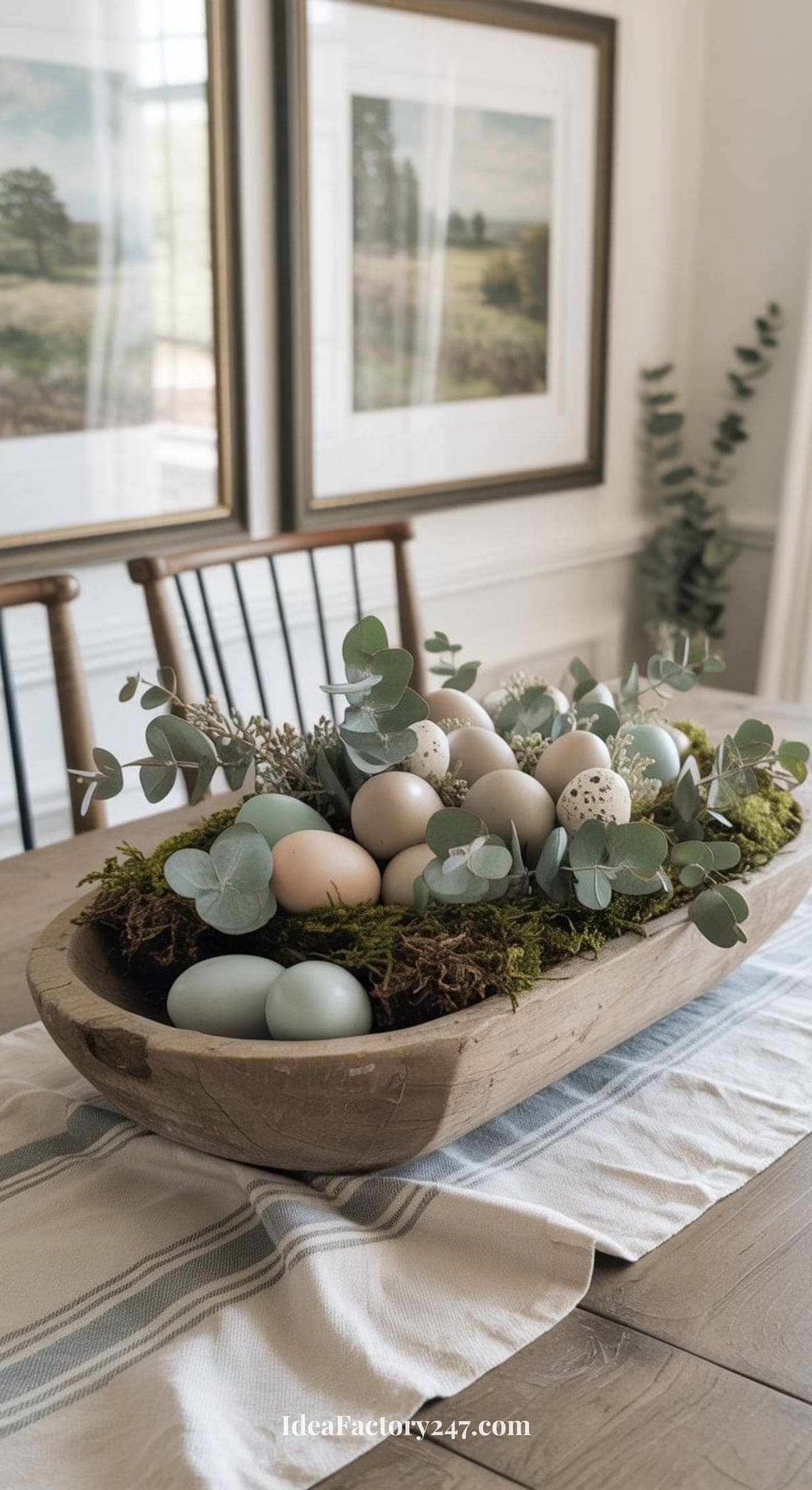 A wooden bowl filled with pastel-colored eggs, moss, and eucalyptus branches serves as a centerpiece on a dining table, with framed landscape art and chairs in the background.