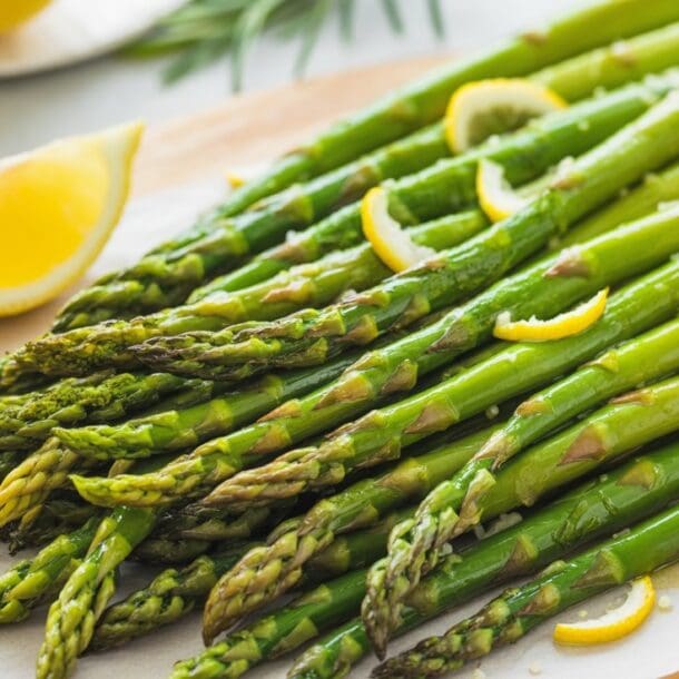 A bunch of cooked asparagus spears garnished with lemon slices and coarse salt, arranged on a parchment-lined wooden board with a lemon wedge nearby.