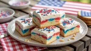 Four square layered cake bars with white frosting and red, white, and blue sprinkles on a plate, with an American flag and a red-checkered napkin in the background.