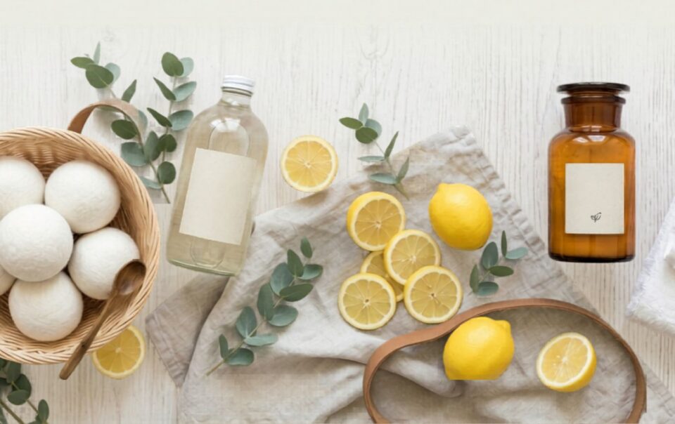 A flat lay of lemons, lemon slices, a glass bottle, a brown jar, white towels, eucalyptus branches, and a basket of white dryer balls with a wooden spoon on a light wooden surface.