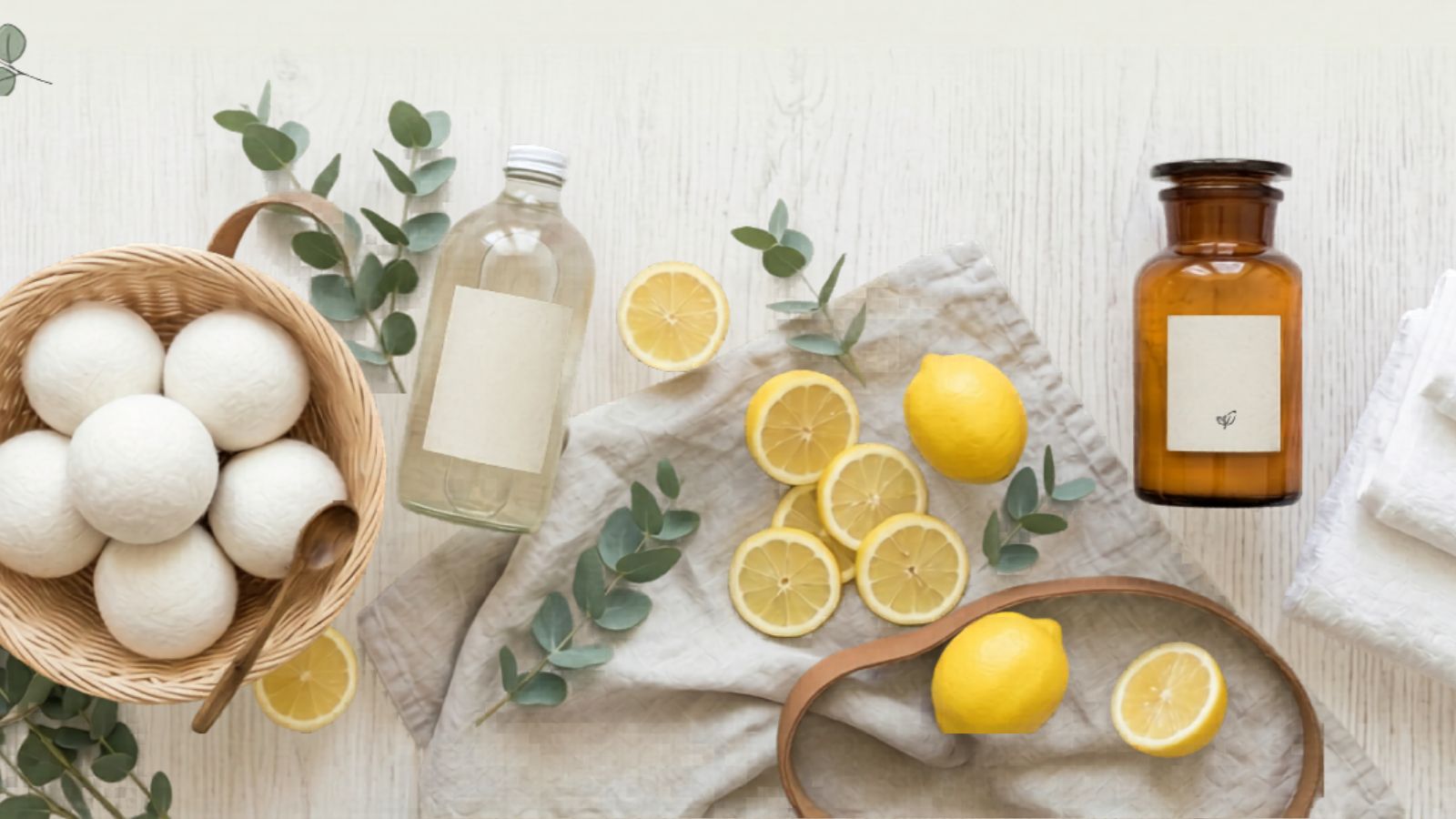 A flat lay of lemons, lemon slices, a glass bottle, a brown jar, white towels, eucalyptus branches, and a basket of white dryer balls with a wooden spoon on a light wooden surface.