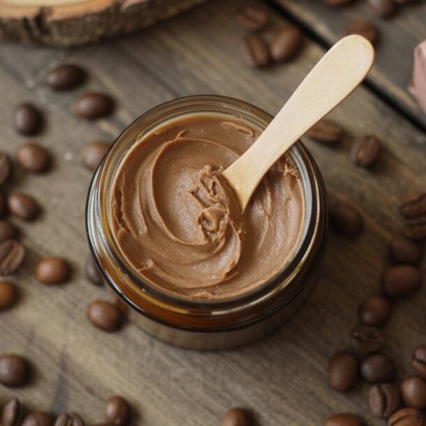 A small jar of creamy brown coffee body butter with a wooden spatula, surrounded by coffee beans on a rustic wooden surface, next to a wrapped gift box with a satin ribbon.