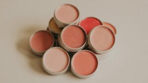 A collection of round metal tins filled with tinted lip balm or cream in various shades of pink, peach, and red, arranged in a loose pile on a neutral surface.