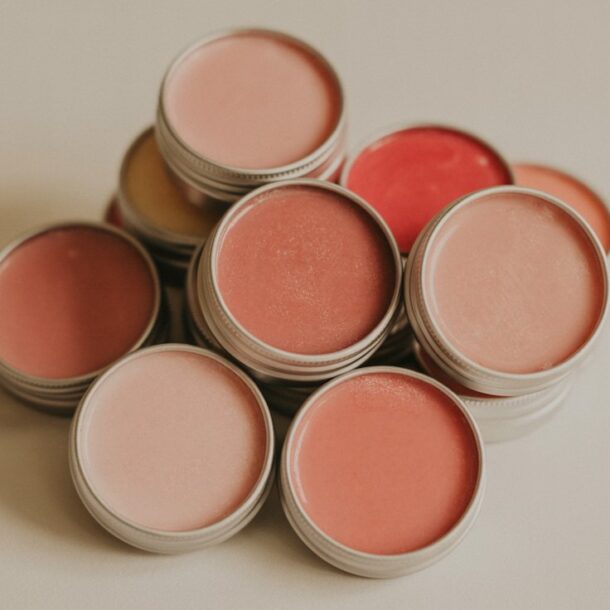 A collection of round metal tins filled with tinted lip balm or cream in various shades of pink, peach, and red, arranged in a loose pile on a neutral surface.