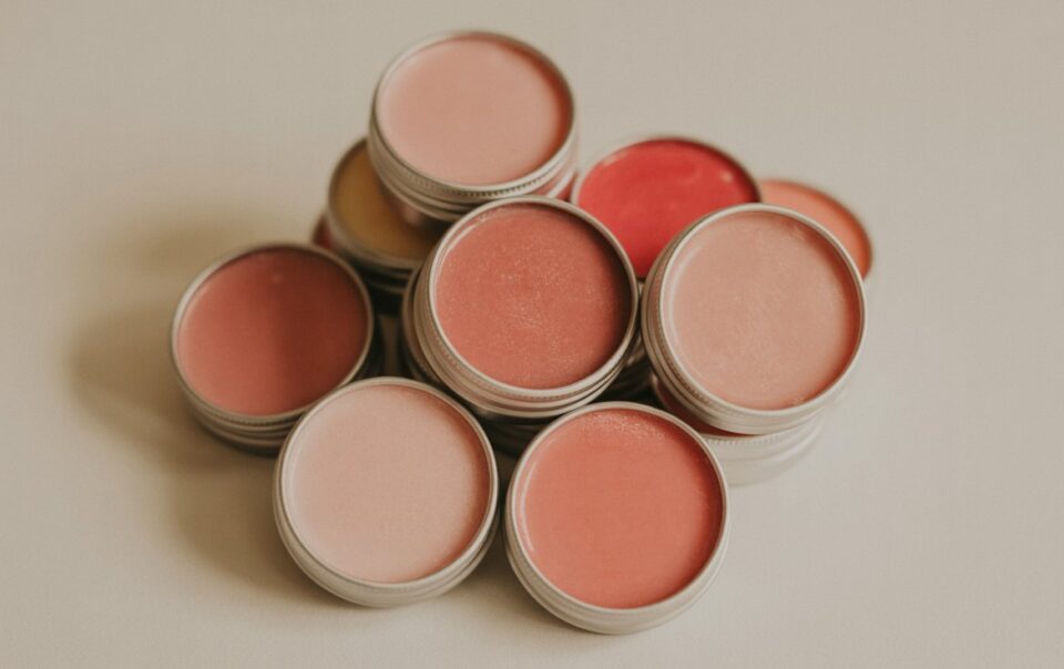 A collection of round metal tins filled with tinted lip balm or cream in various shades of pink, peach, and red, arranged in a loose pile on a neutral surface.