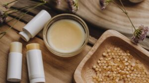 Three tubes of lip balm, a small glass jar of balm, and a wooden tray with yellow beeswax pellets are arranged on a wooden surface with purple flowers—perfect for illustrating how to make lip balm at home.