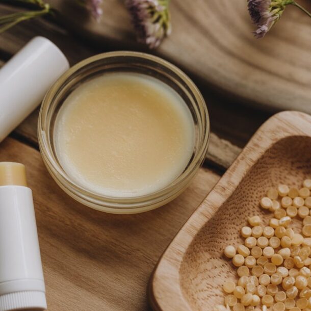 Three tubes of lip balm, a small glass jar of balm, and a wooden tray with yellow beeswax pellets are arranged on a wooden surface with purple flowers—perfect for illustrating how to make lip balm at home.