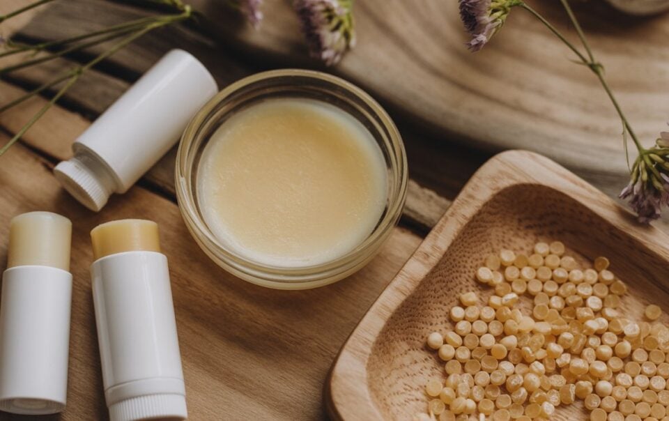 Three tubes of lip balm, a small glass jar of balm, and a wooden tray with yellow beeswax pellets are arranged on a wooden surface with purple flowers—perfect for illustrating how to make lip balm at home.