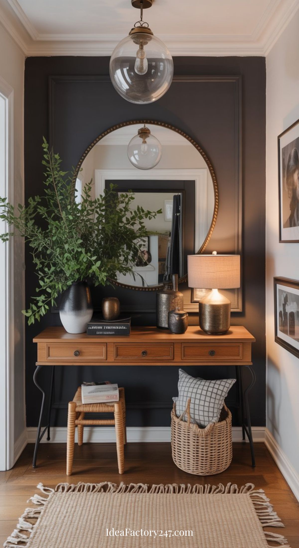 A stylish entryway features a wooden console table with drawers, topped with a lamp, books, vases, and greenery. A round mirror hangs above, with framed photos on the wall and a basket with pillows on the floor.