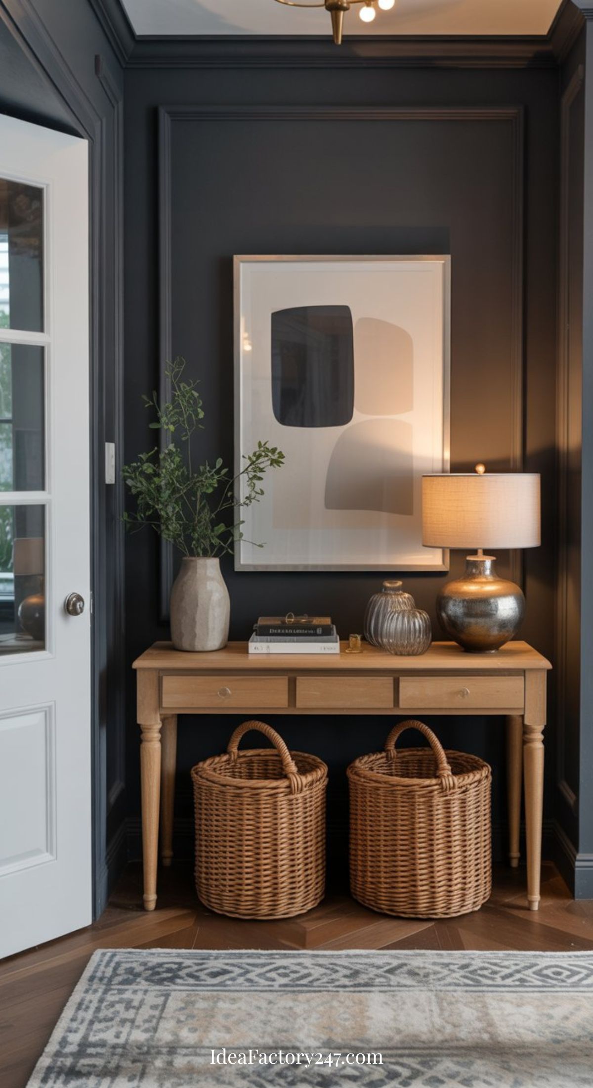 A wooden console table with two wicker baskets underneath, a table lamp, vases, and books on top, sits against a dark-paneled wall featuring abstract art. A door and a rug are partially visible.