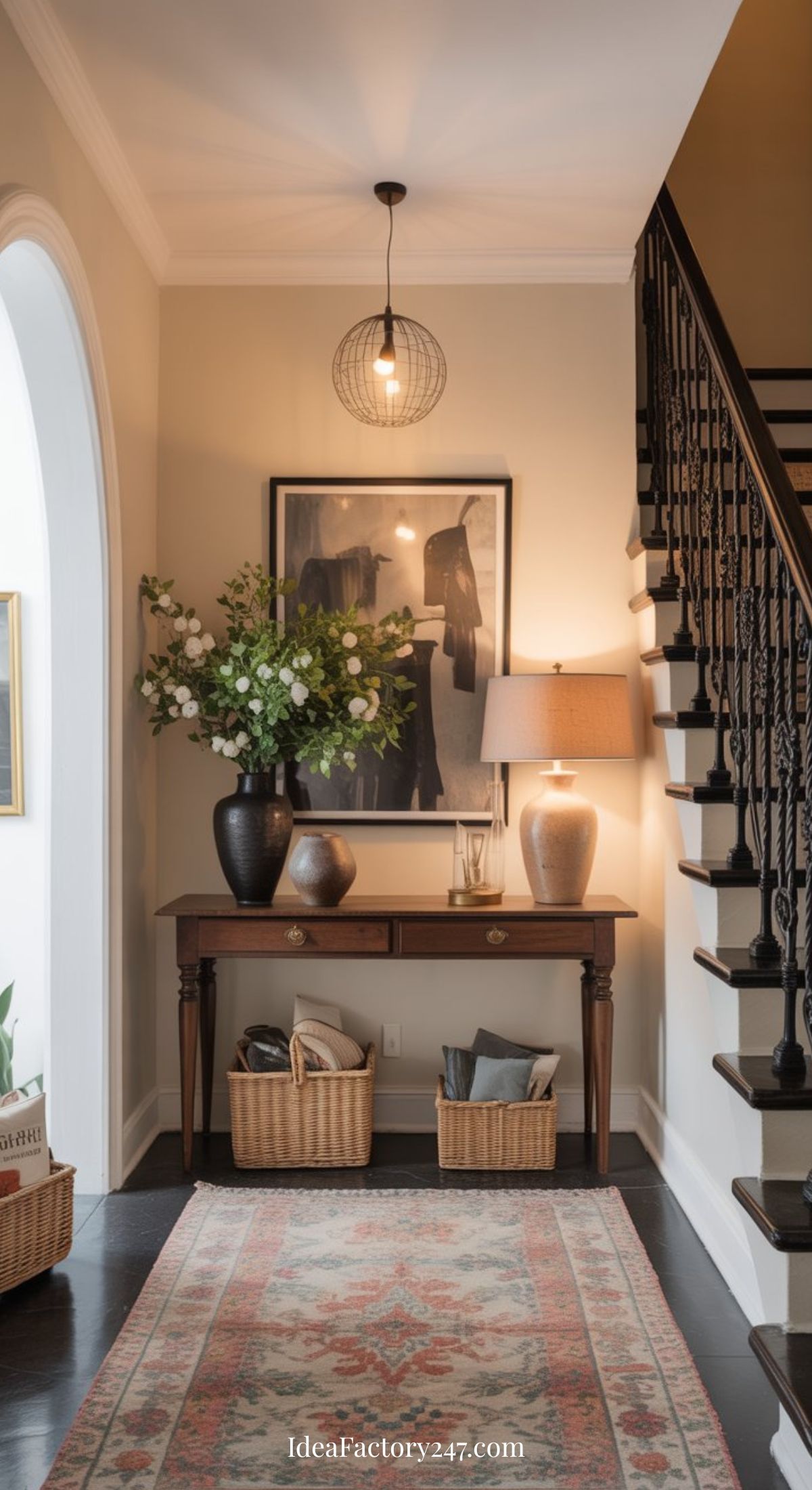 A stylish hallway features a Nancy Meyers entryway table topped with a lamp, white flowers, and decor. Two baskets rest below, while an ornate rug, framed art, and a black-railed staircase complete the cozy scene.